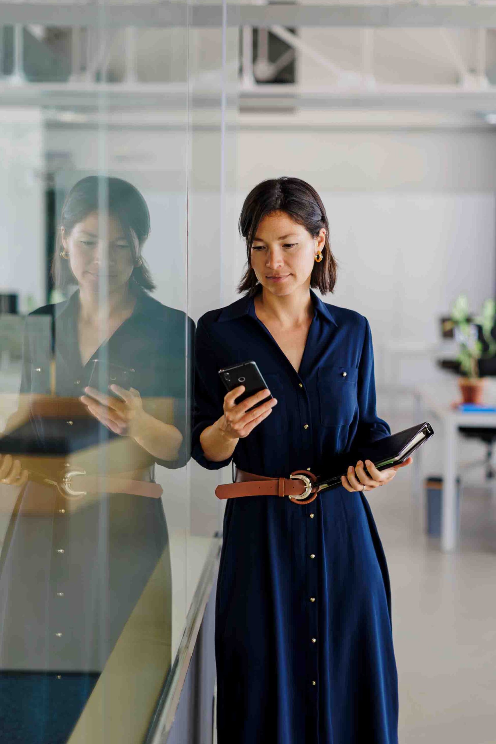 A Woman with a blue dress holding a smartphone leaning on a office glass wall