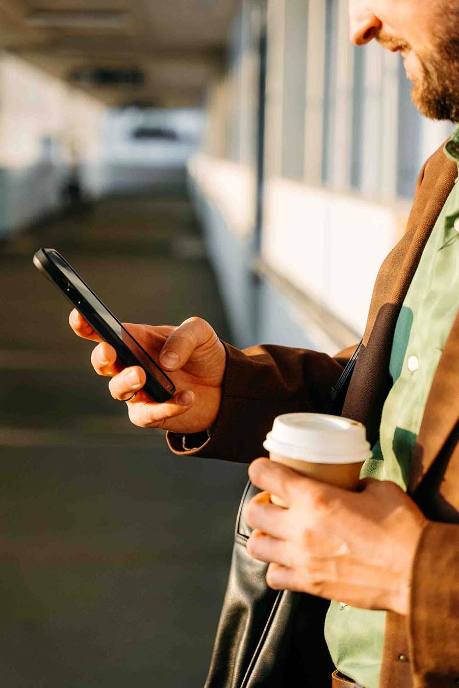 A men holding and looking to his phone and holding a cup of coffee to go somwhere outside